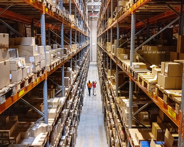 Gut sortiertes Hochregallager mit vielen Paketen und zwei Mitarbeitern