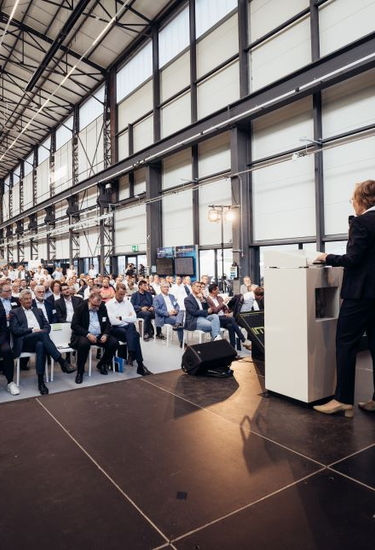 Frau steht auf einer Bühne der Smarten Demonstrationsfabrik in Siegen und spricht zum Publikum